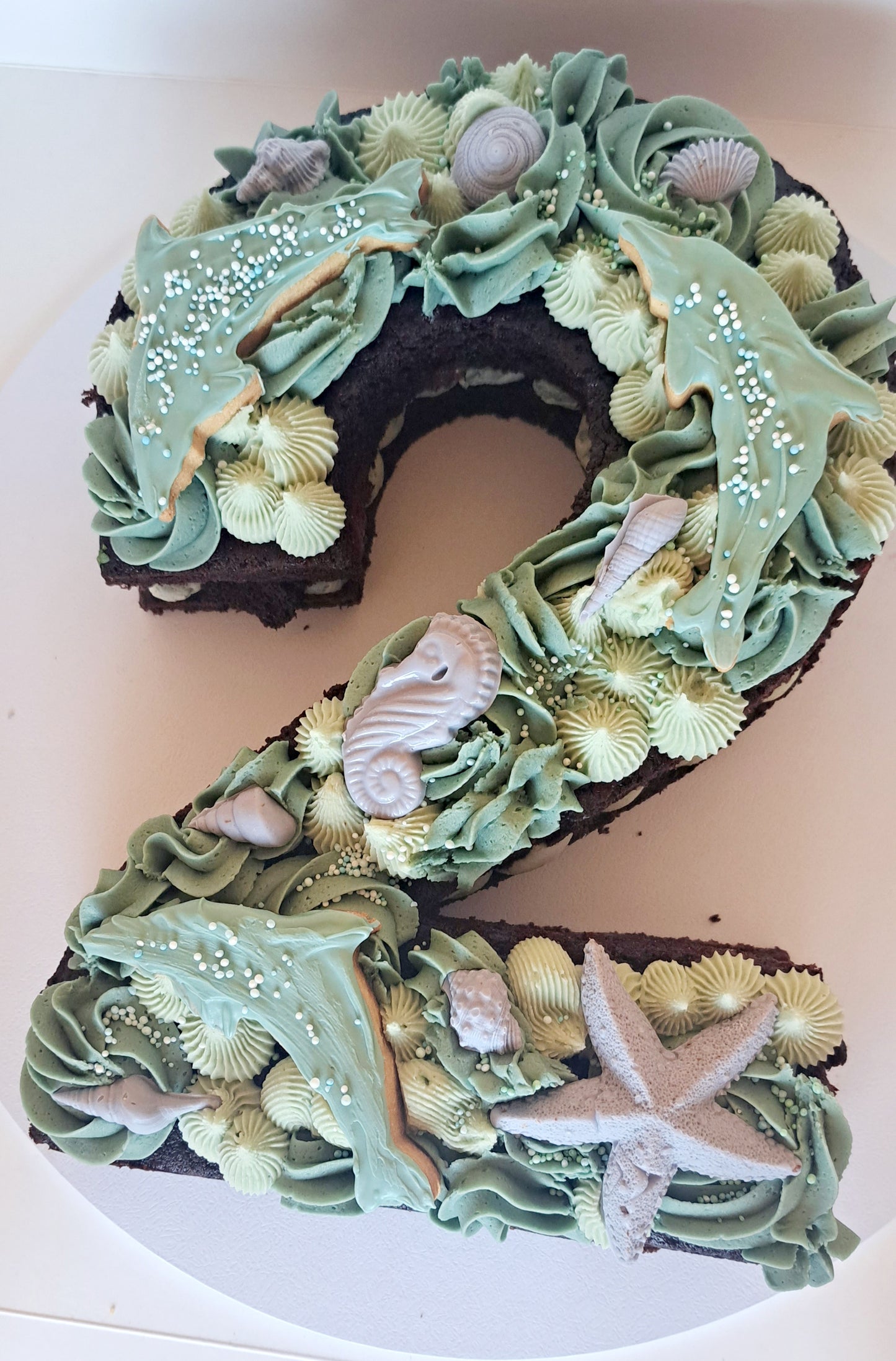 A chocolate cake in the shape of the number 2, topped with ocean green buttercream swirls, dolphin biscuits and sea animal chocolates.  